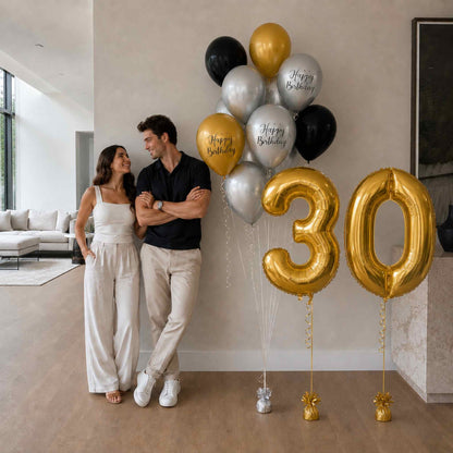 Couple standing next to large '30' gold balloons in a modern living room.