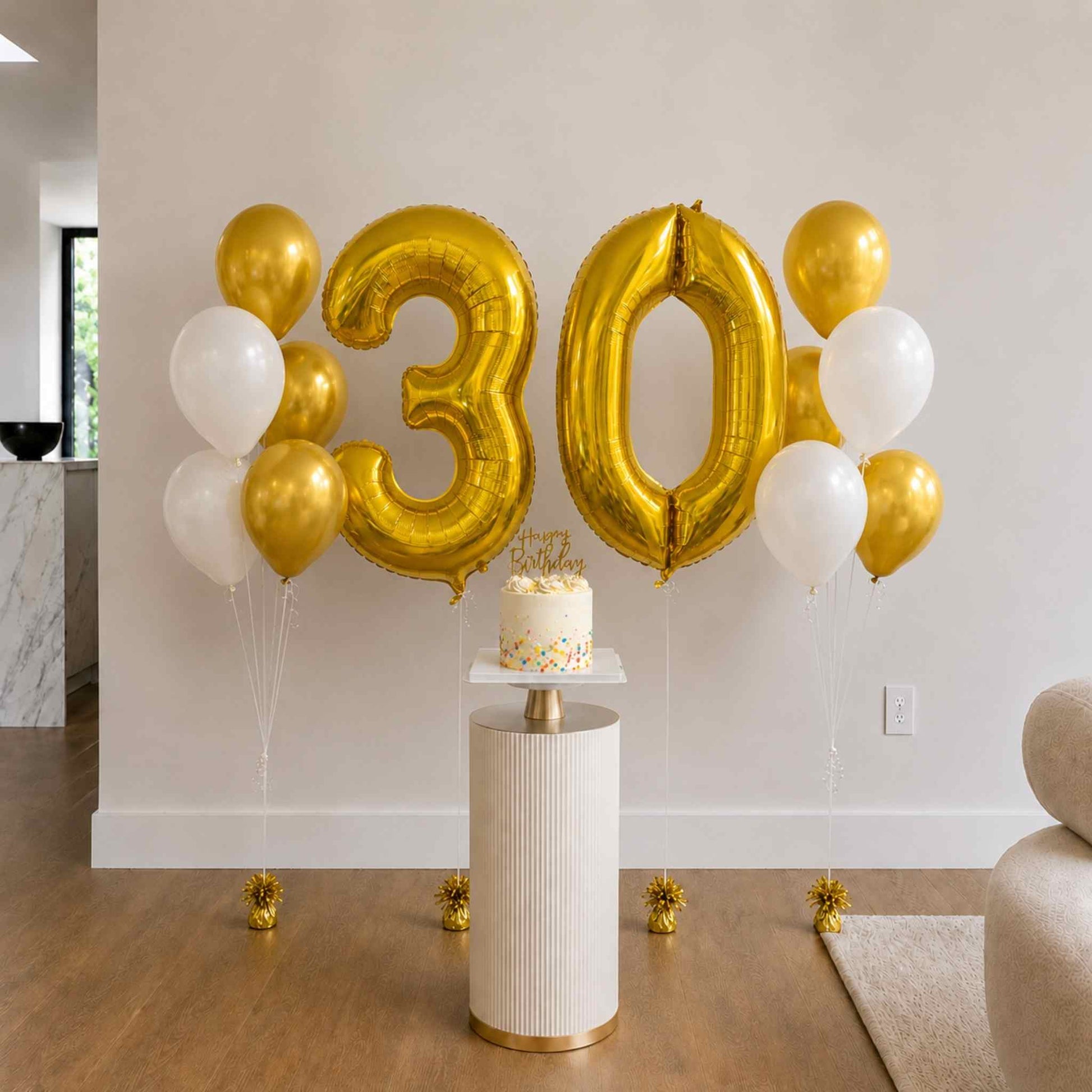 Gold '30' balloons and a small cake on a white pedestal in a room.