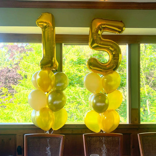 Gold and yellow number balloons spelling '15' in front of a window with greenery outside.