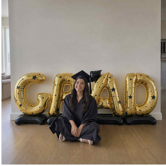 Graduate in cap and gown sitting on the floor with 'GRAD' balloons spelling out 'GRAD' in a room.