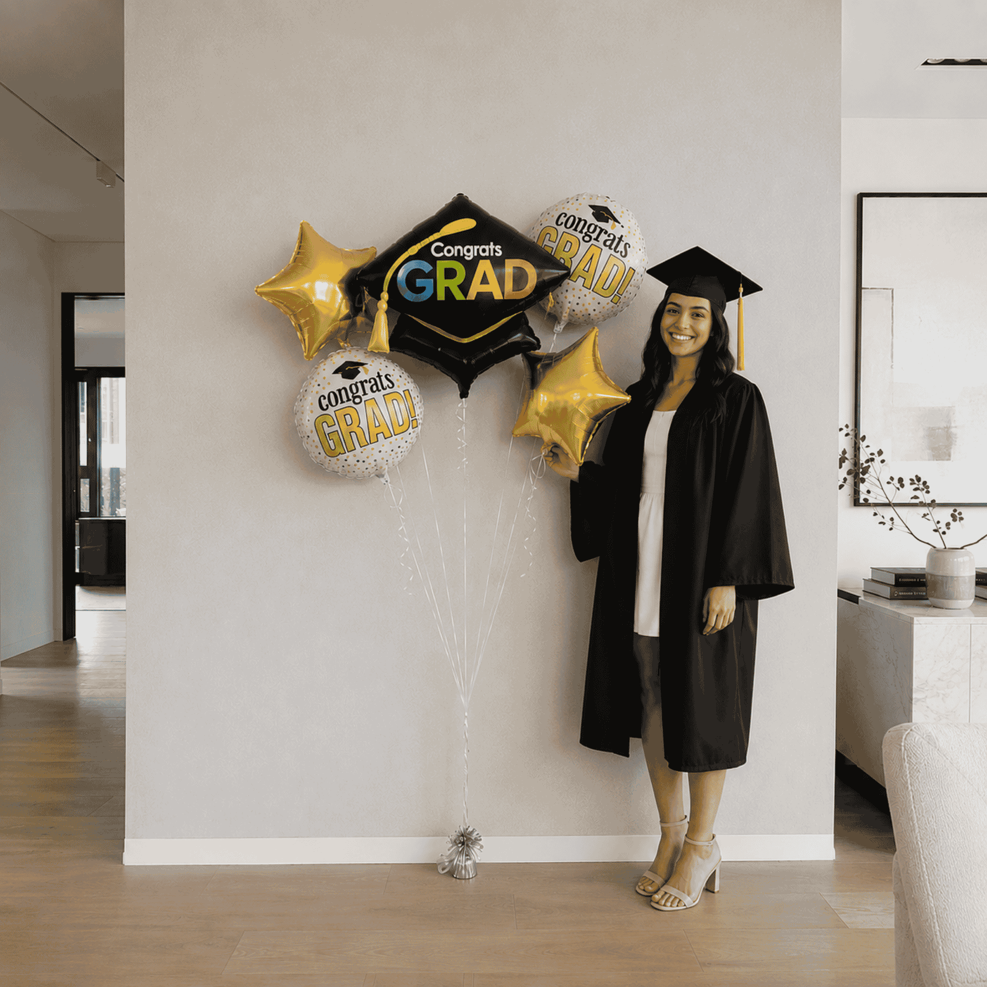 Graduate in cap and gown standing next to graduation balloons in a home setting.