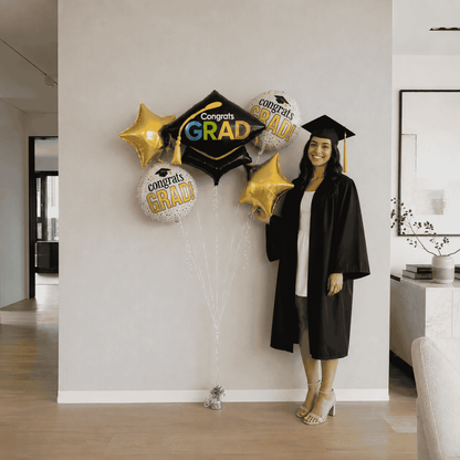 Graduate in cap and gown standing next to graduation balloons in a home setting.