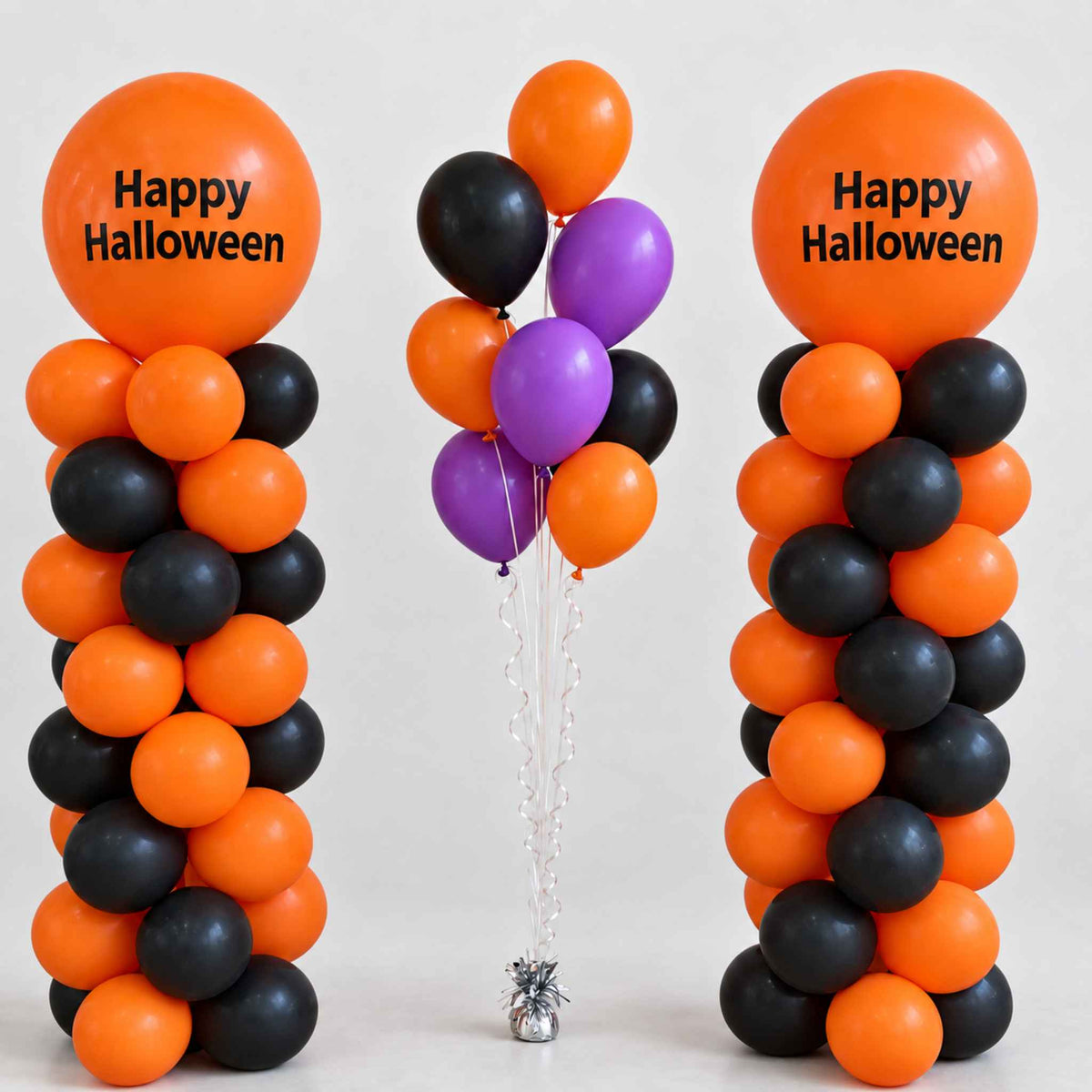 Halloween balloon decoration set with two tall columns of orange and black balloons, each topped with a large orange balloon printed 'Happy Halloween,' plus a central cluster of orange, black, and purple balloons tied together on a clear stand.