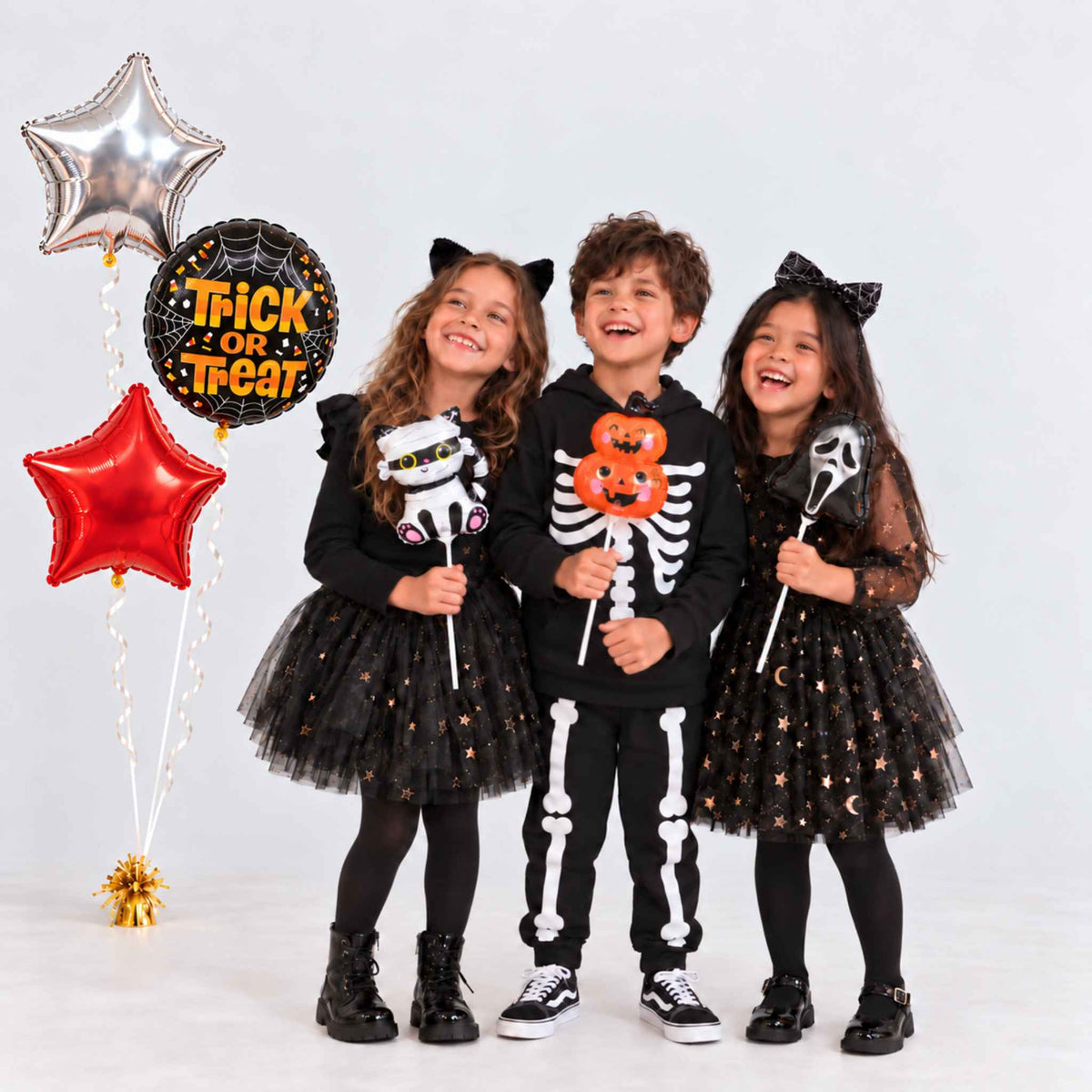 Three children wearing Halloween costumes pose with decorative balloons on a light gray studio background. The child on the left wears a black dress with gold stars and a black top, holding a white lollipop.