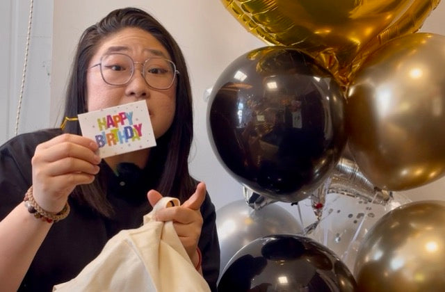 A woman wearing glasses holds up a small white card that says 'HAPPY BIRTHDAY' in colorful letters. She is holding a cream-colored fabric pouch in her other hand, with black and gold balloons in the background.