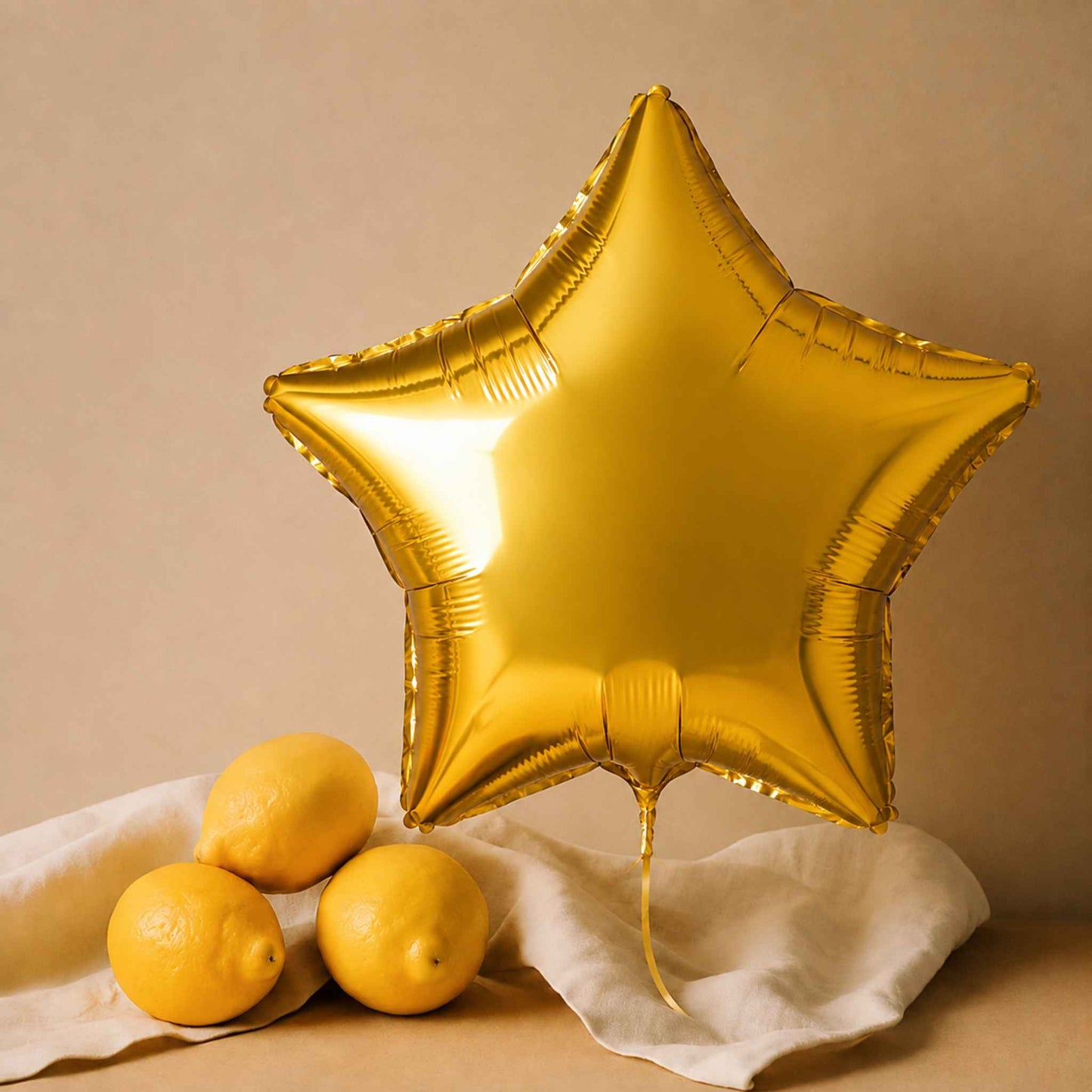 A metallic gold star-shaped foil balloon stands upright on a draped white cloth beside three whole lemons against a warm beige background.