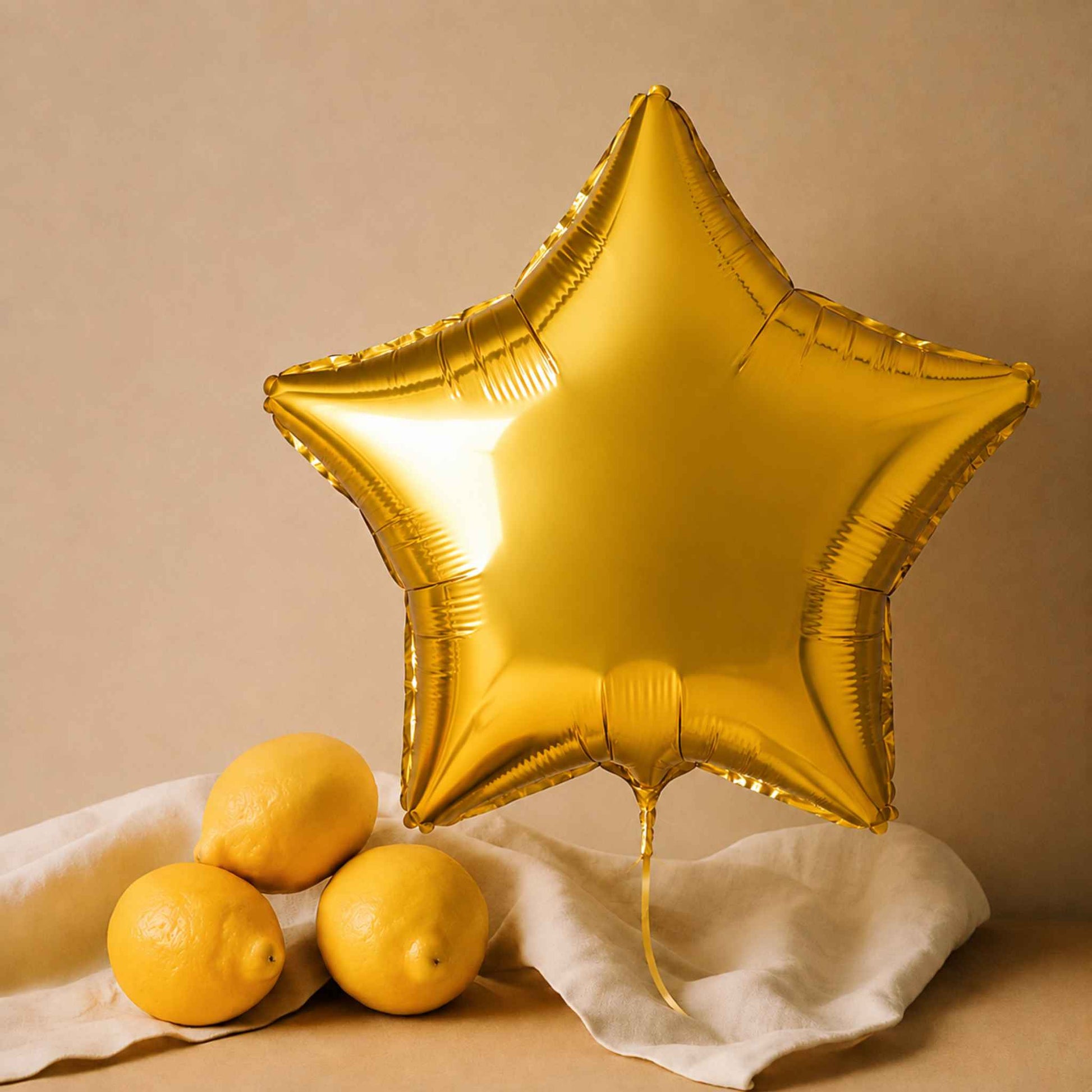 A metallic gold star-shaped foil balloon stands upright on a draped white cloth beside three whole lemons against a warm beige background.