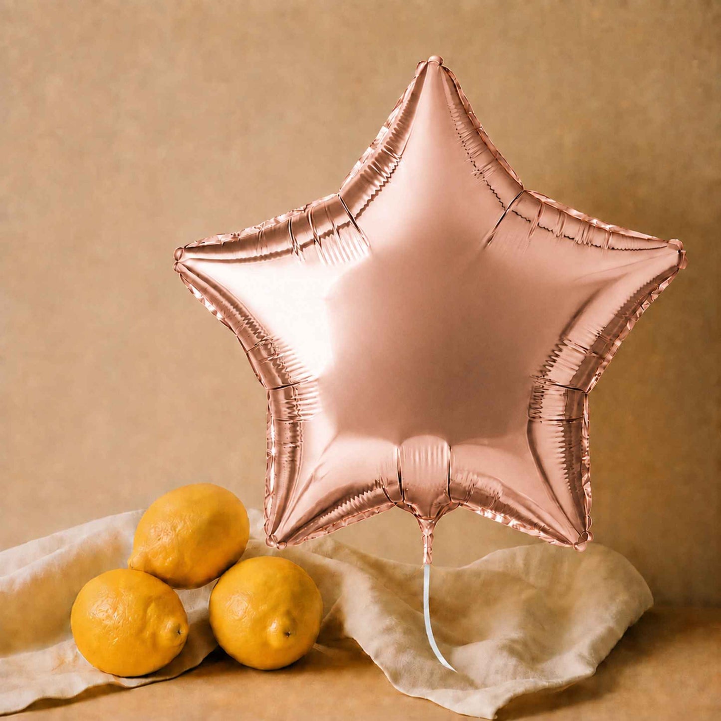 A large metallic rose gold star-shaped foil balloon floating above three yellow lemons on a beige cloth against a warm beige background.