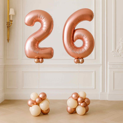 Rose gold helium number balloons “2” and “6” displayed on cream and rose gold balloon bases in an elegant white room.