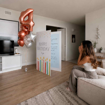 A woman sits on a sofa in a living room, looking surprised and happy as a large birthday card and metallic number 4 balloon stand nearby. The birthday setup includes a colorful candle-themed card, a handwritten message card, and rose gold, silver, and white balloons.