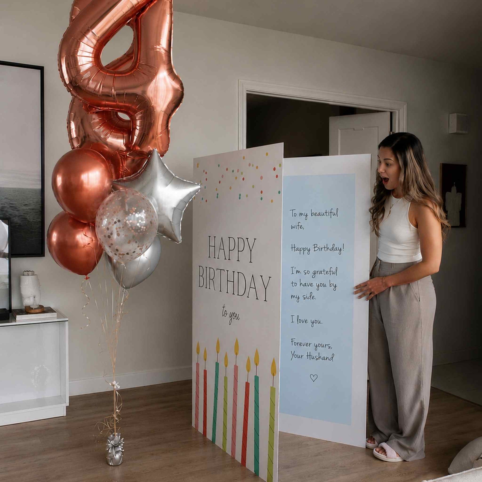 A woman stands beside a large birthday card in a home interior, with a rose-gold balloon shaped like the number 4, metallic balloons, and a candle-themed 'Happy Birthday' sign on the floor.