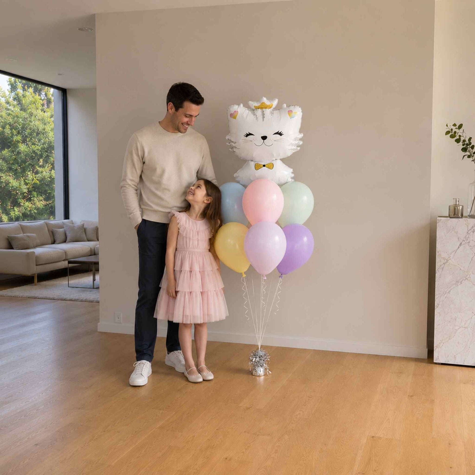 Man and young girl standing in a living room with a large cat-shaped balloon and pastel balloons.