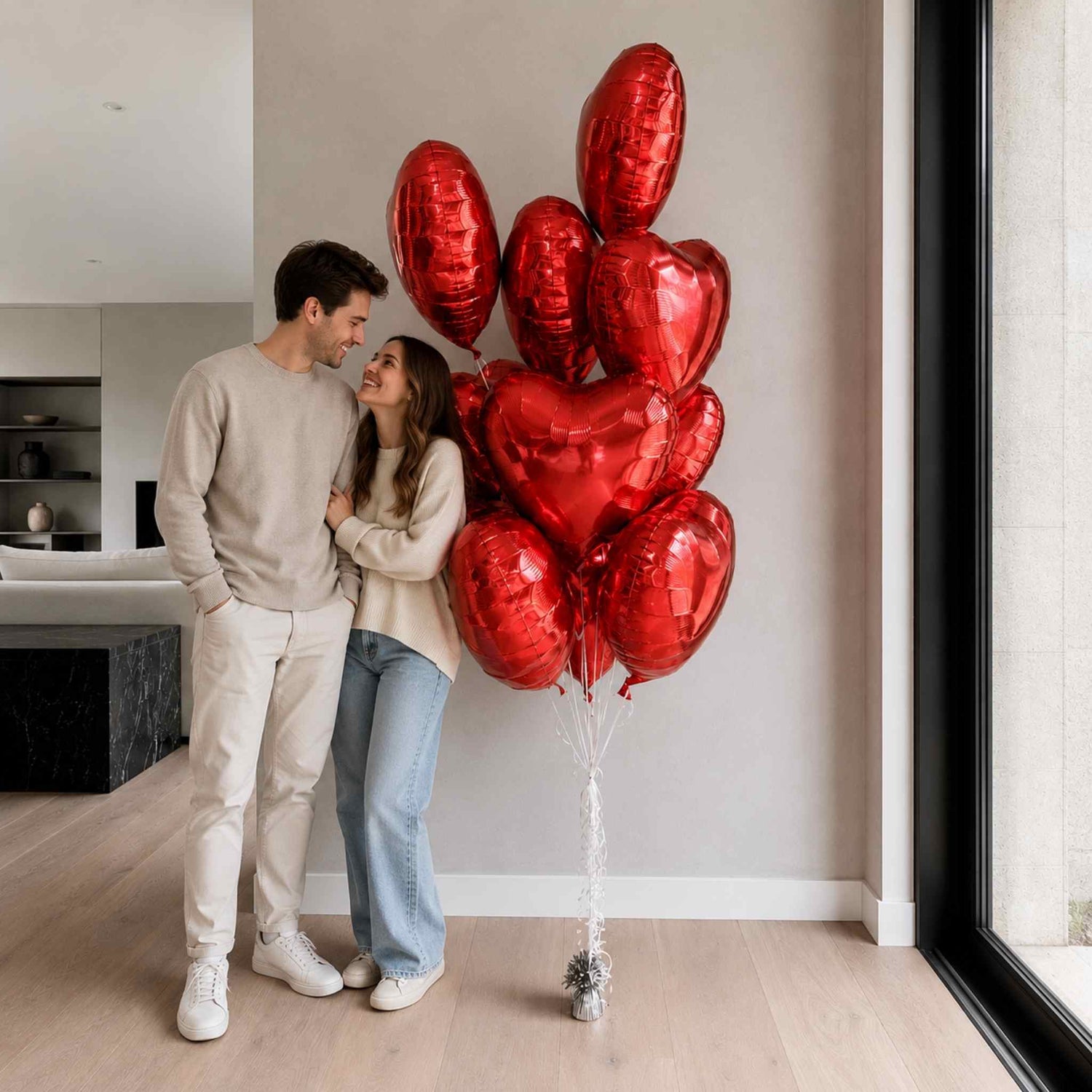 Couple standing in a modern living room with red heart-shaped balloons.