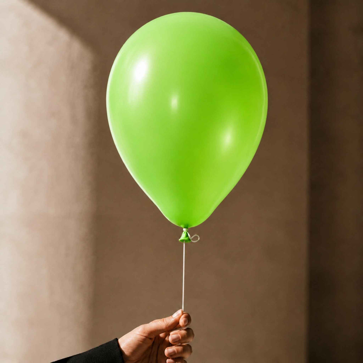 A bright lime green latex balloon held by a hand against a warm, textured brown background.