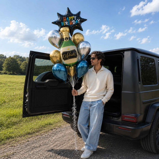 A man in a cream half-zip sweater and light blue jeans stands beside an open matte black SUV, holding a personalized balloon bouquet with a star-shaped 'Happy Birthday' balloon, gold and silver balloons, and a champagne bottle balloon.