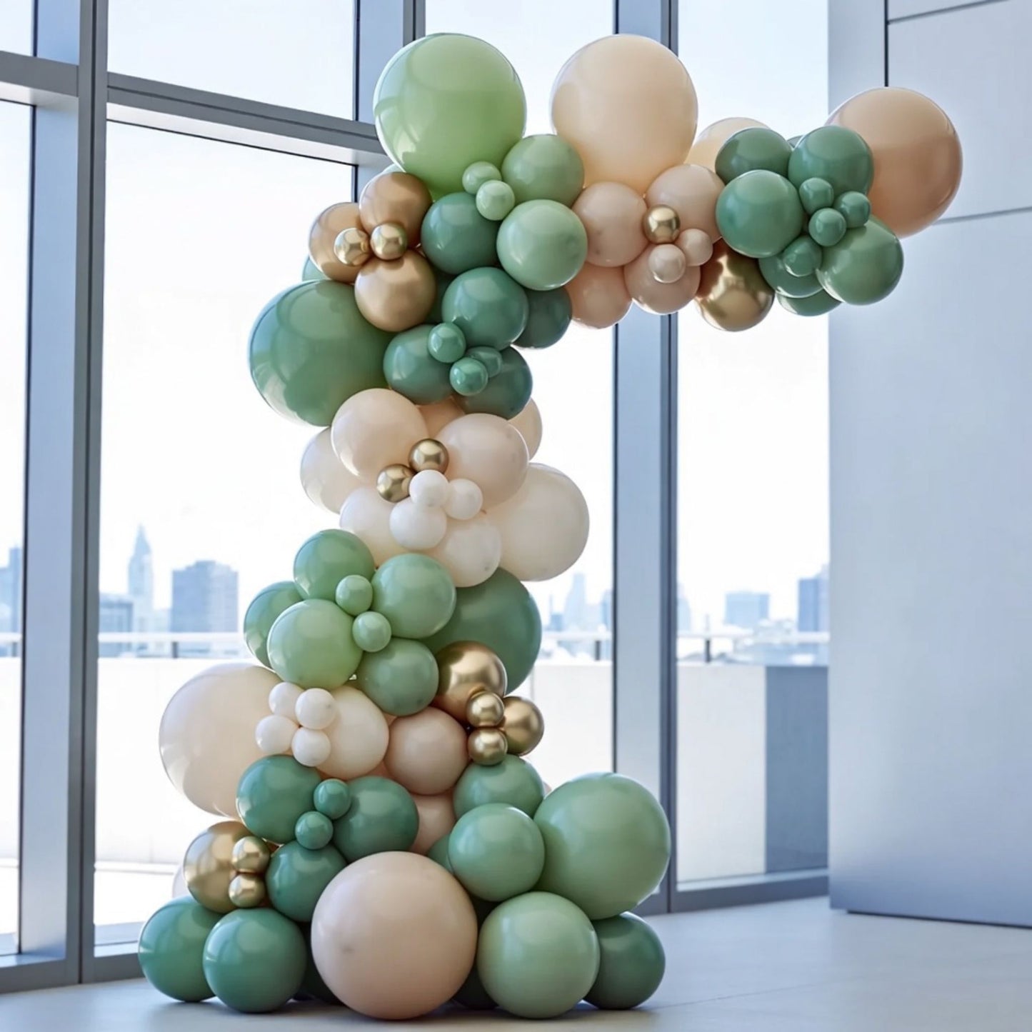Balloon arch garland with green, beige, and gold balloons in a room with large windows.