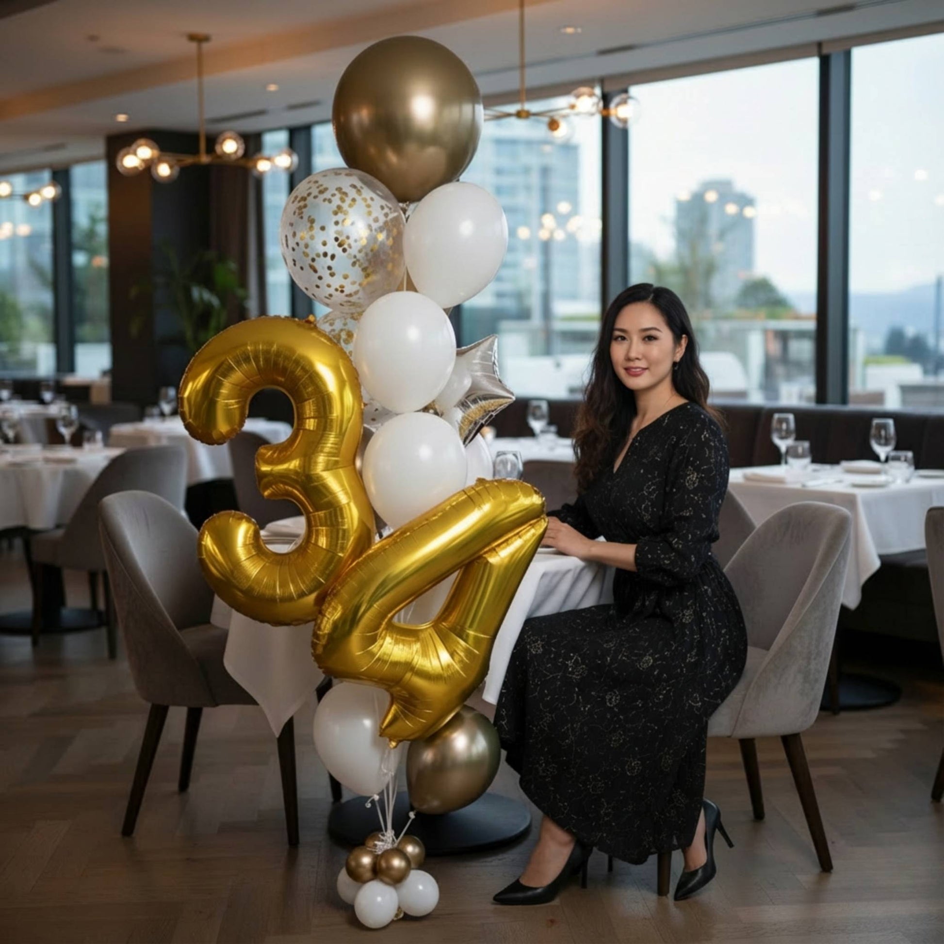 Woman sitting at a table with large gold and white balloons shaped like the number 34 in a restaurant setting.