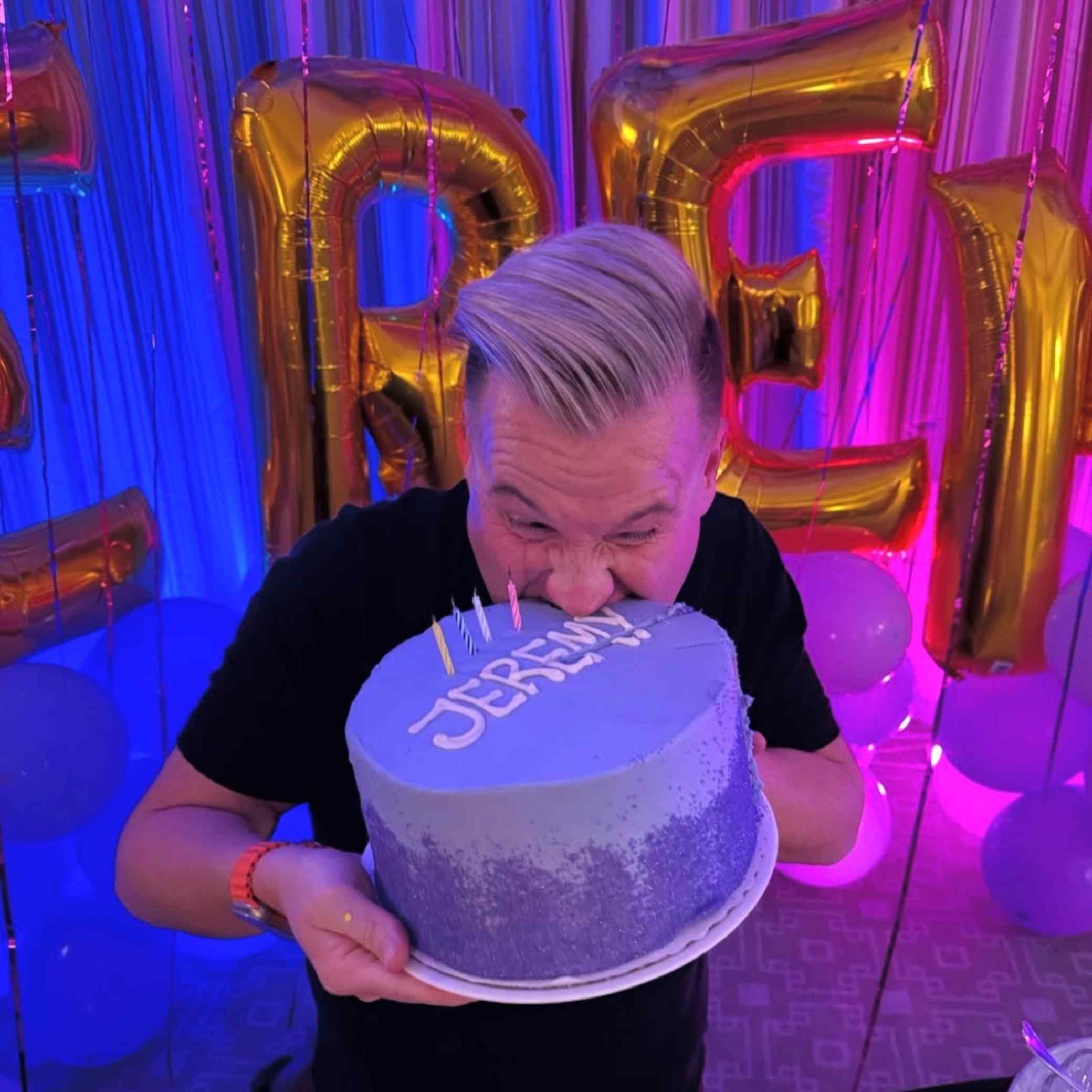 Person blowing out candles on a cake with 'Jeremy' written on it, surrounded by colorful balloons and decorations.