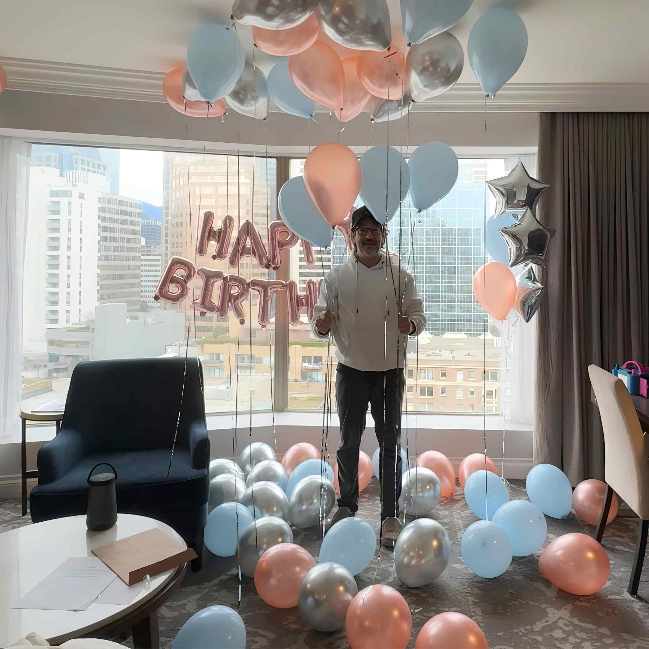 Man standing in a room with colorful balloons and 'Happy Birthday' sign.