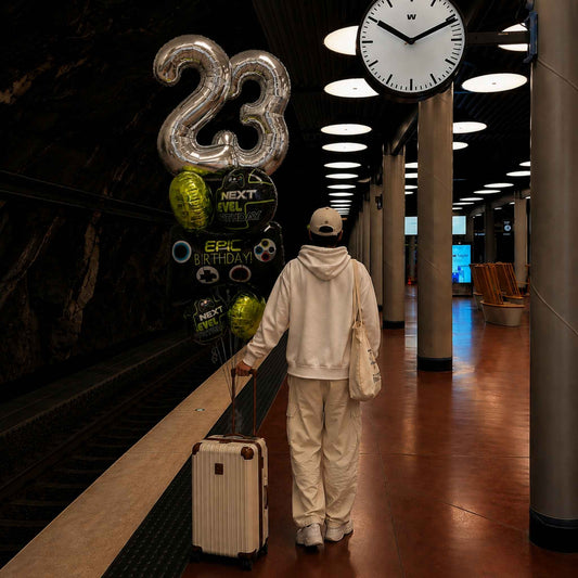 A person in a matching cream-colored hoodie and sweatpants walks away on a train platform, pulling a silver suitcase and holding a cluster of birthday balloons that spell '23' and say 'Next Level Epic Birthday'.