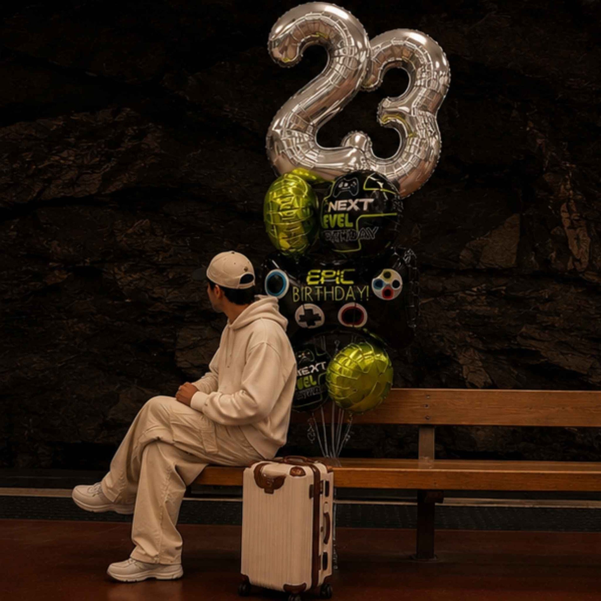 A person in a beige tracksuit sits on a wooden bench next to a large birthday balloon bouquet shaped like the number 23, with a small cream suitcase on the floor in front. The background is a dark textured wall.