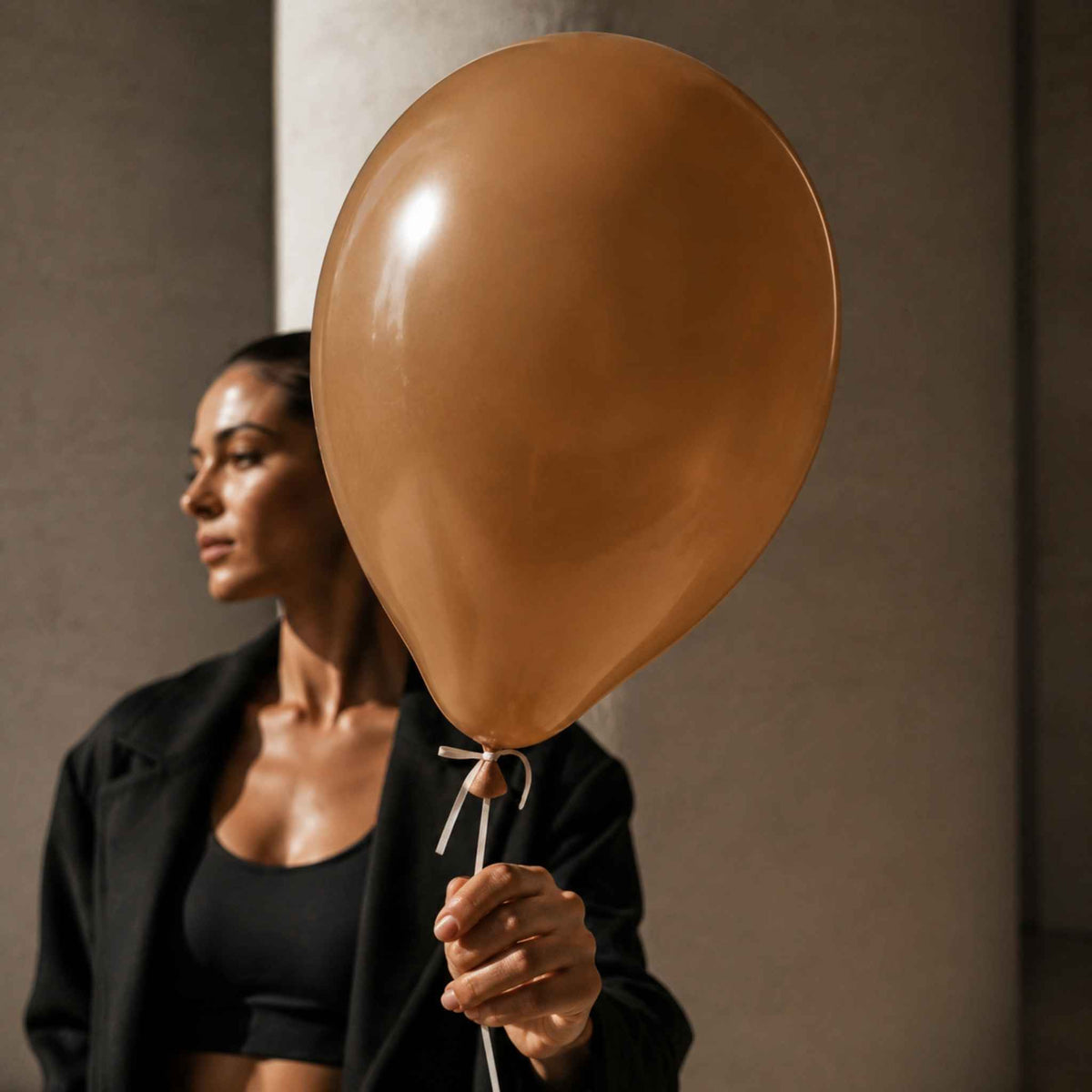 A woman in a black blazer and black sports bra holds a large, glossy, metallic bronze balloon in front of her face, set against a minimalist textured gray wall.