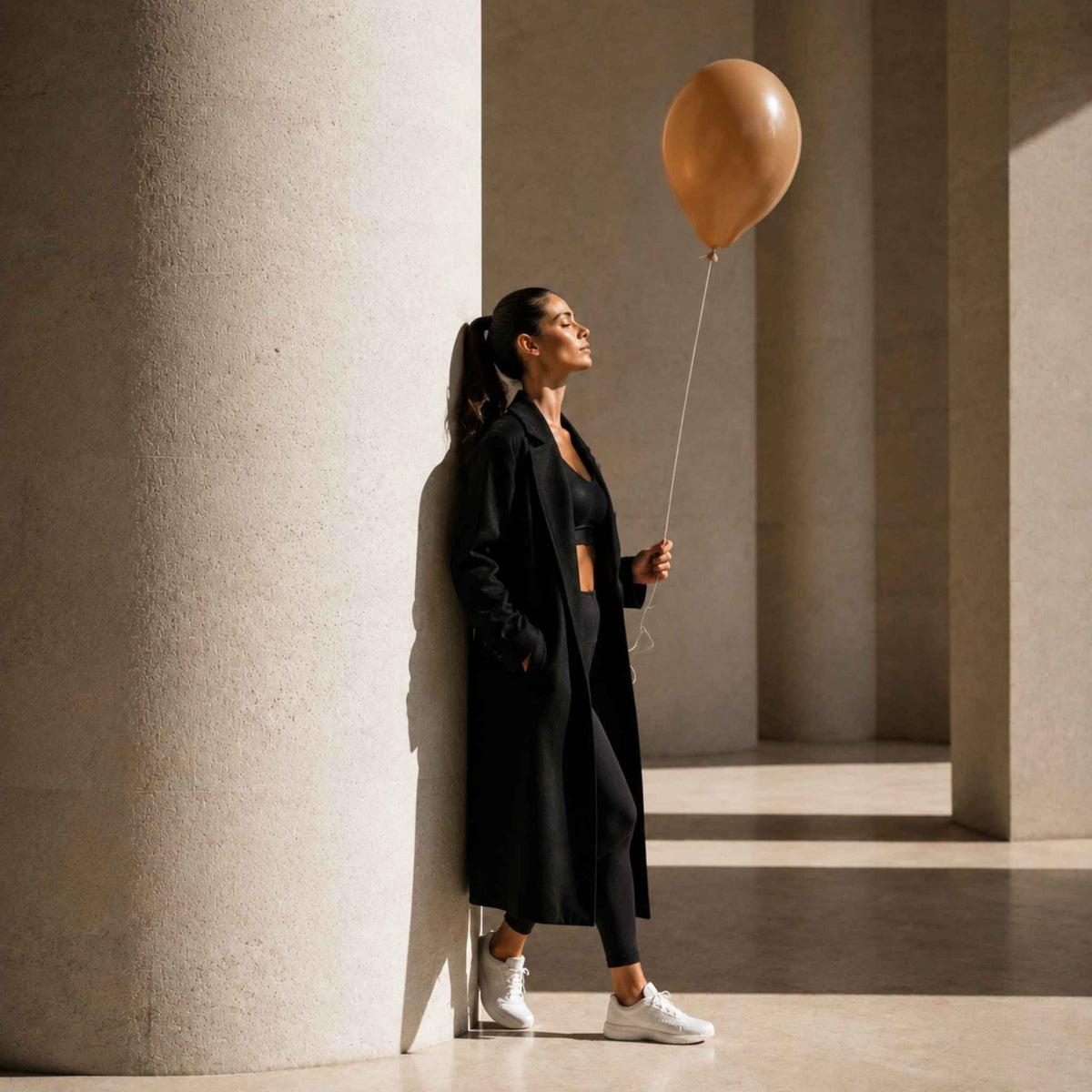 A woman in a long black coat, black top and pants, and white sneakers stands beside a large concrete column holding a tan balloon in a sunlit architectural corridor.