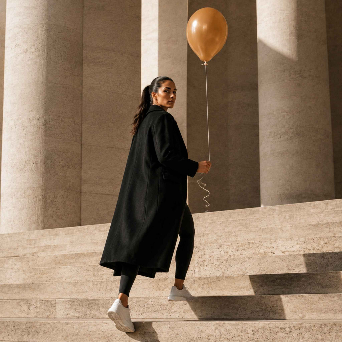 A woman in a long black coat, black leggings, and white sneakers walks down stone steps while holding a gold balloon, set against a background of large beige columns.