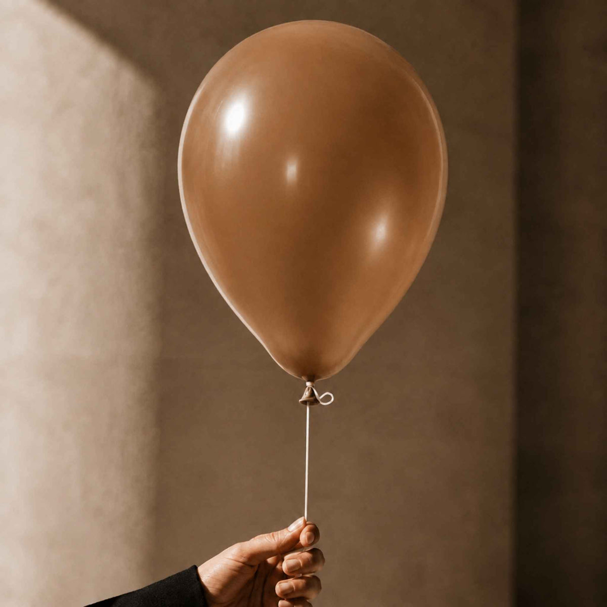 A matte taupe-brown latex balloon held by a hand against a warm, textured beige background.
