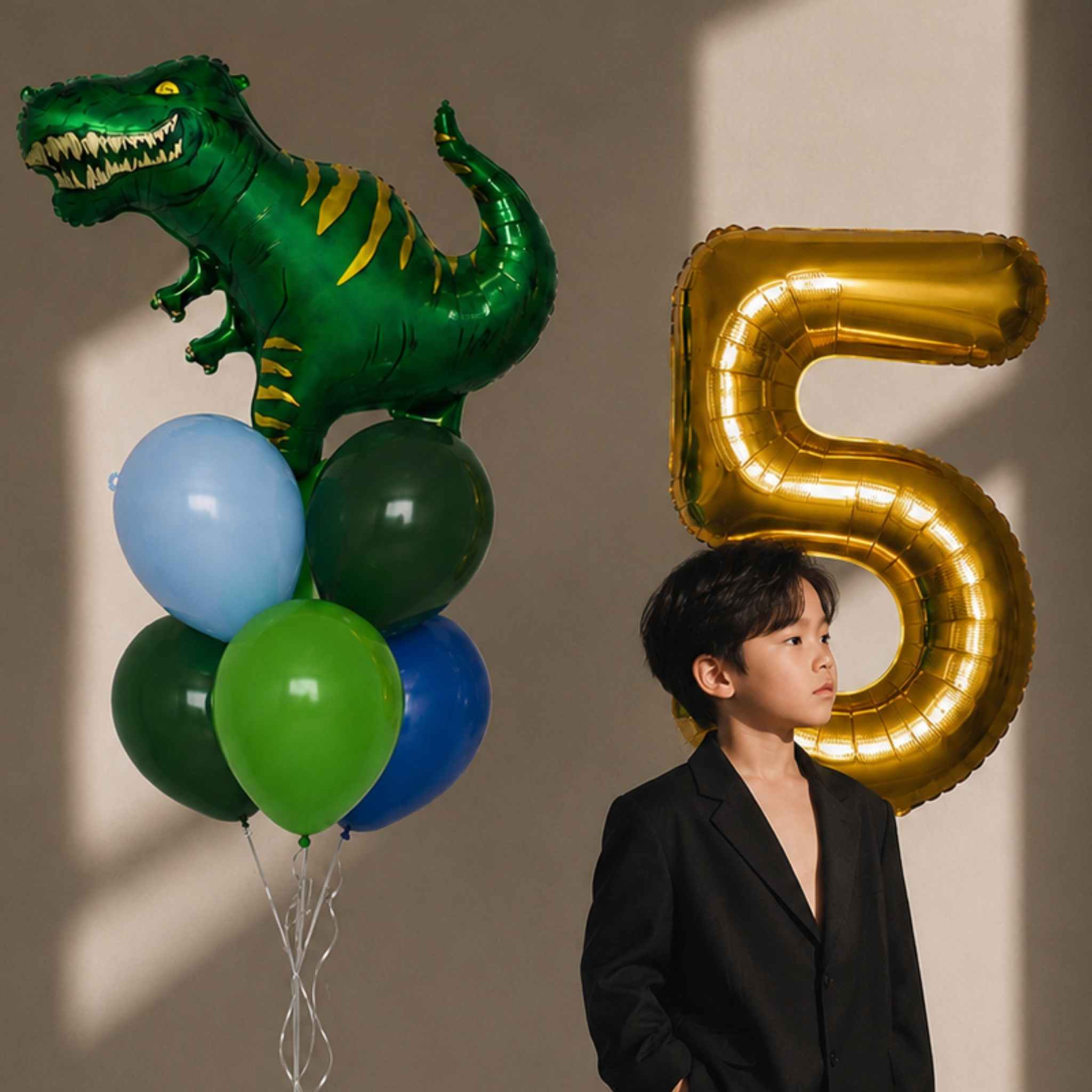 A child in a black blazer stands beside a large metallic gold balloon number 5 and a green dinosaur balloon with yellow stripes, alongside smaller blue, green, and dark balloons, against a beige wall with sunlight shadows.