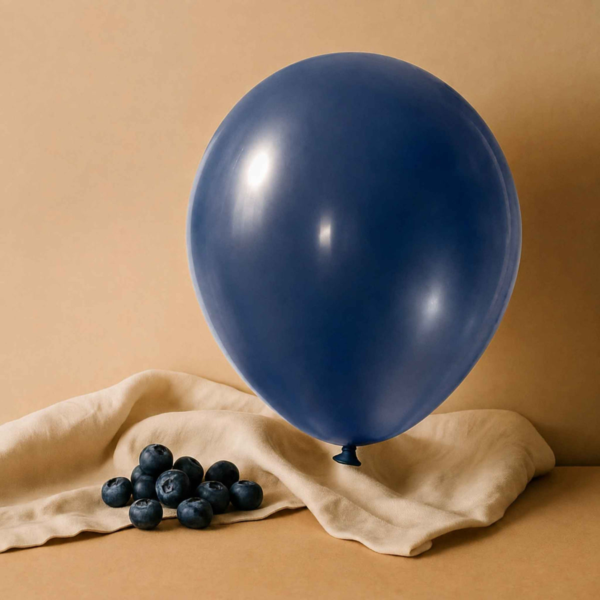 A single matte navy blue balloon stands upright on draped beige fabric with a small cluster of blueberries in front, against a warm beige studio background.
