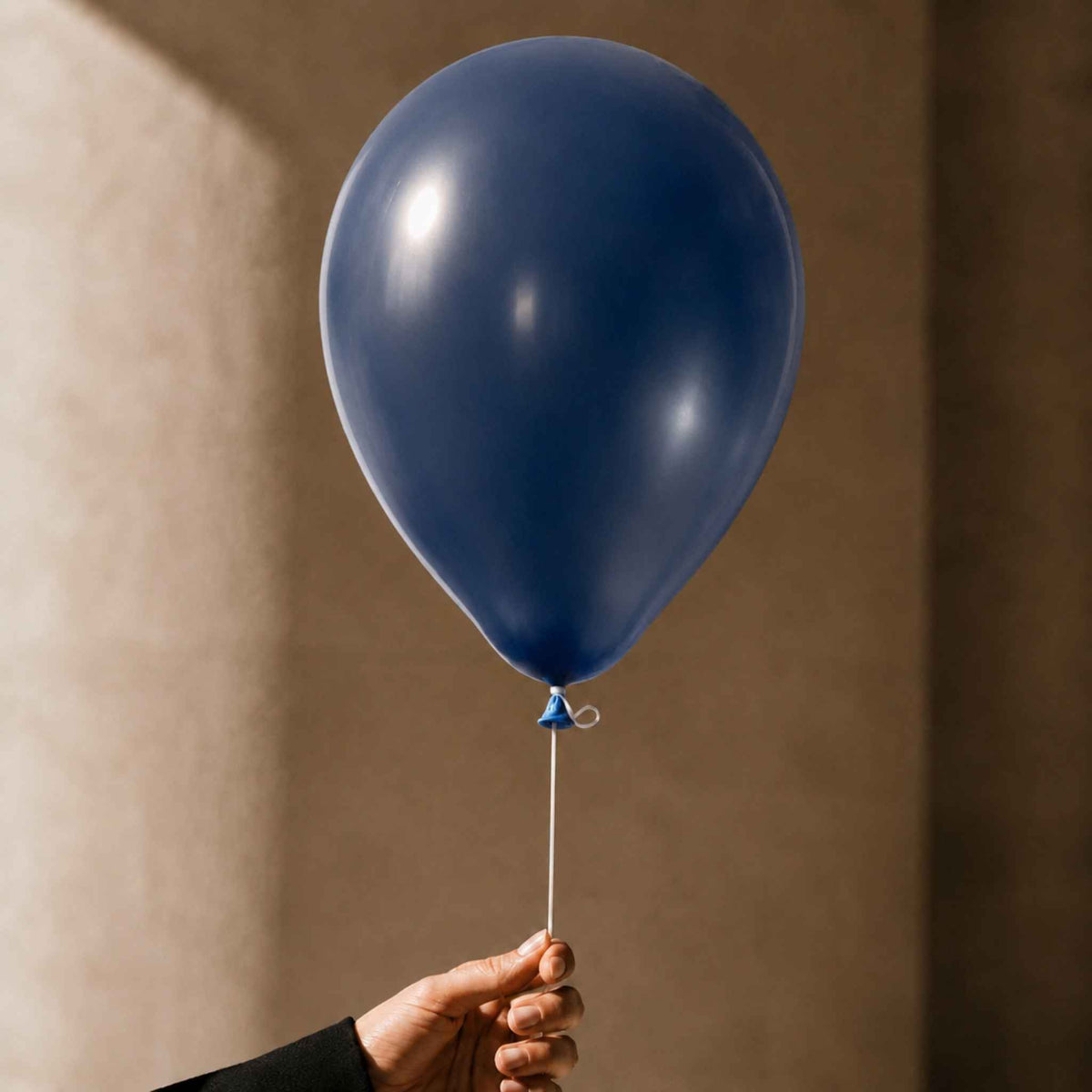 A single matte navy blue balloon held by a hand against a warm beige wall background.
