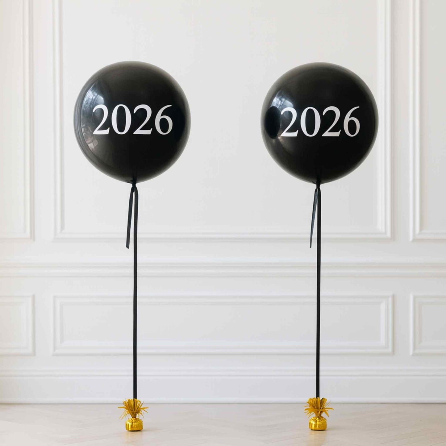 Two black balloons with '2026' printed on them against a white paneled wall.