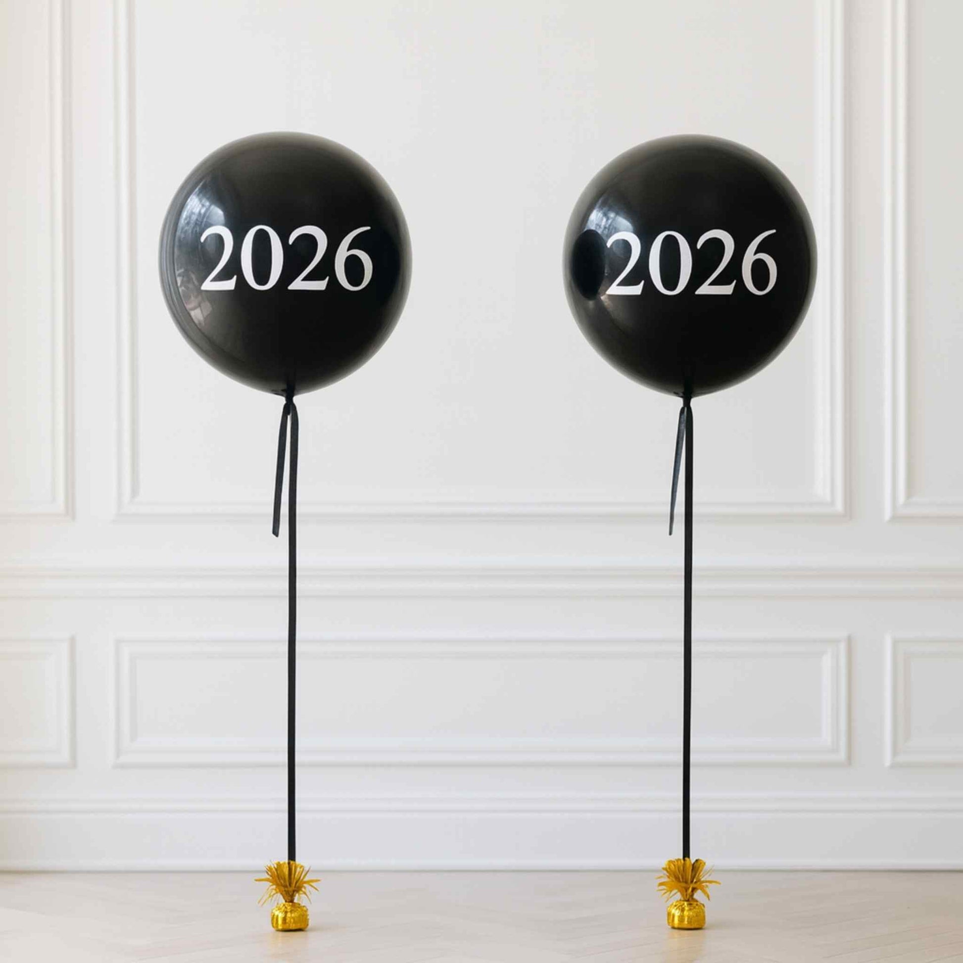 Two black balloons with '2026' printed on them against a white paneled wall.