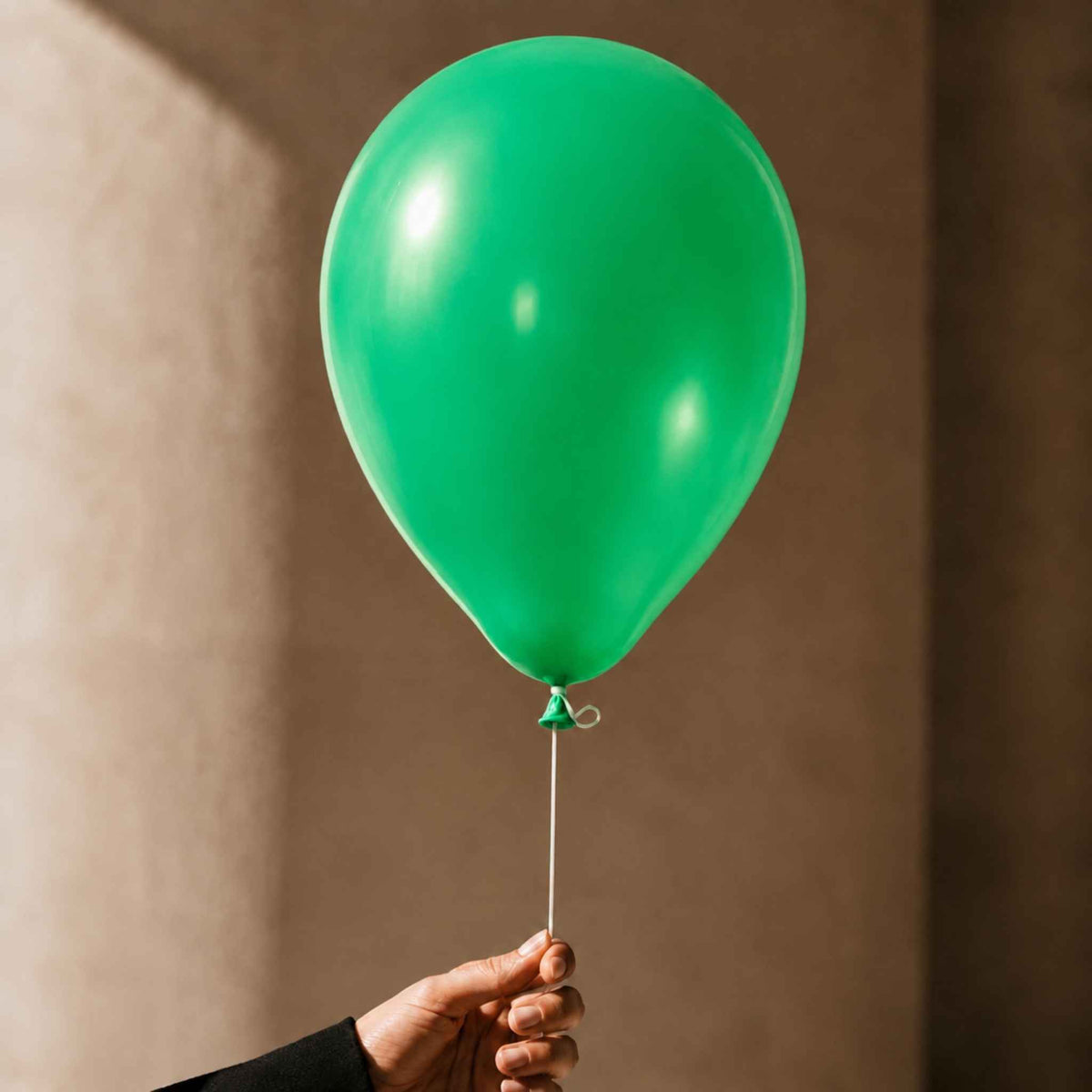 A bright green latex balloon held by a hand against a warm, textured beige wall.