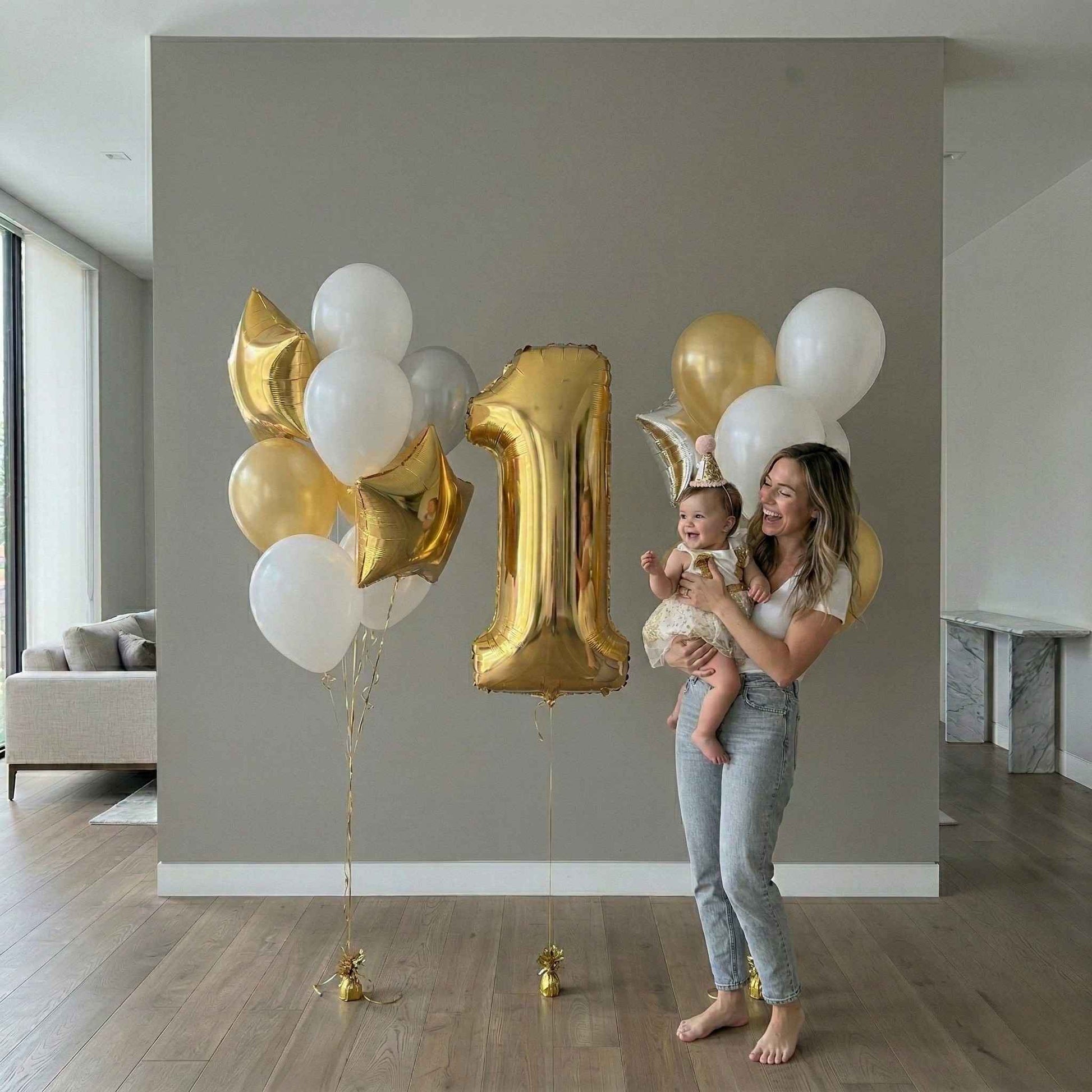 Woman holding a baby with gold and white balloons, including a large '1' shaped balloon, in a modern living room.