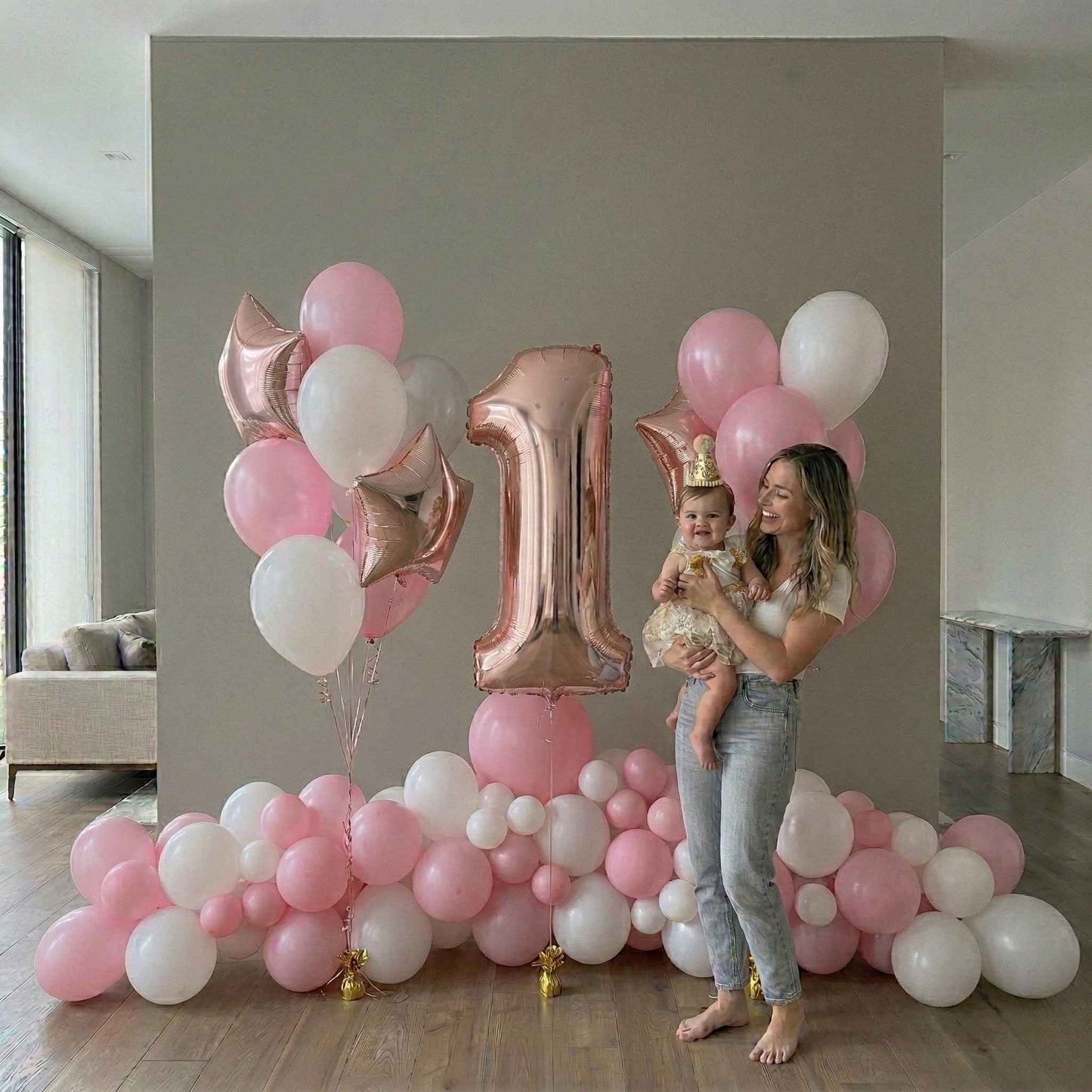 Woman holding a baby in front of a pink and white balloon arrangement with a large number '1' balloon.