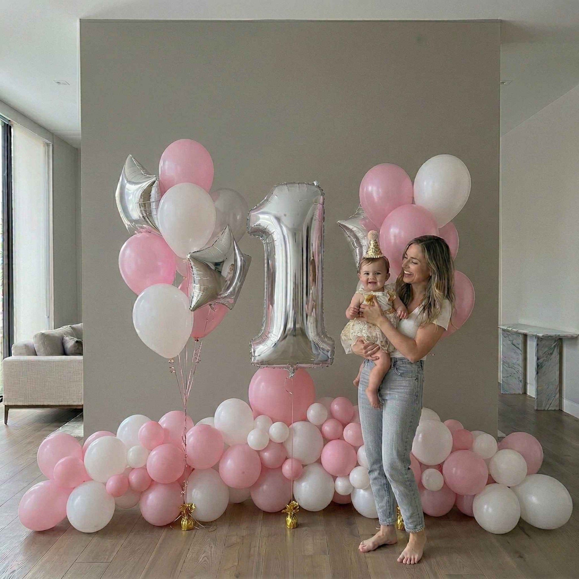 Woman holding a baby in front of a large '1' balloon with pink and white balloons in a room.