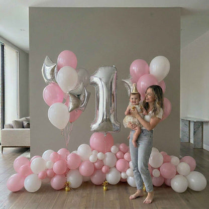 Woman holding a baby in front of a large '1' balloon with pink and white balloons in a room.