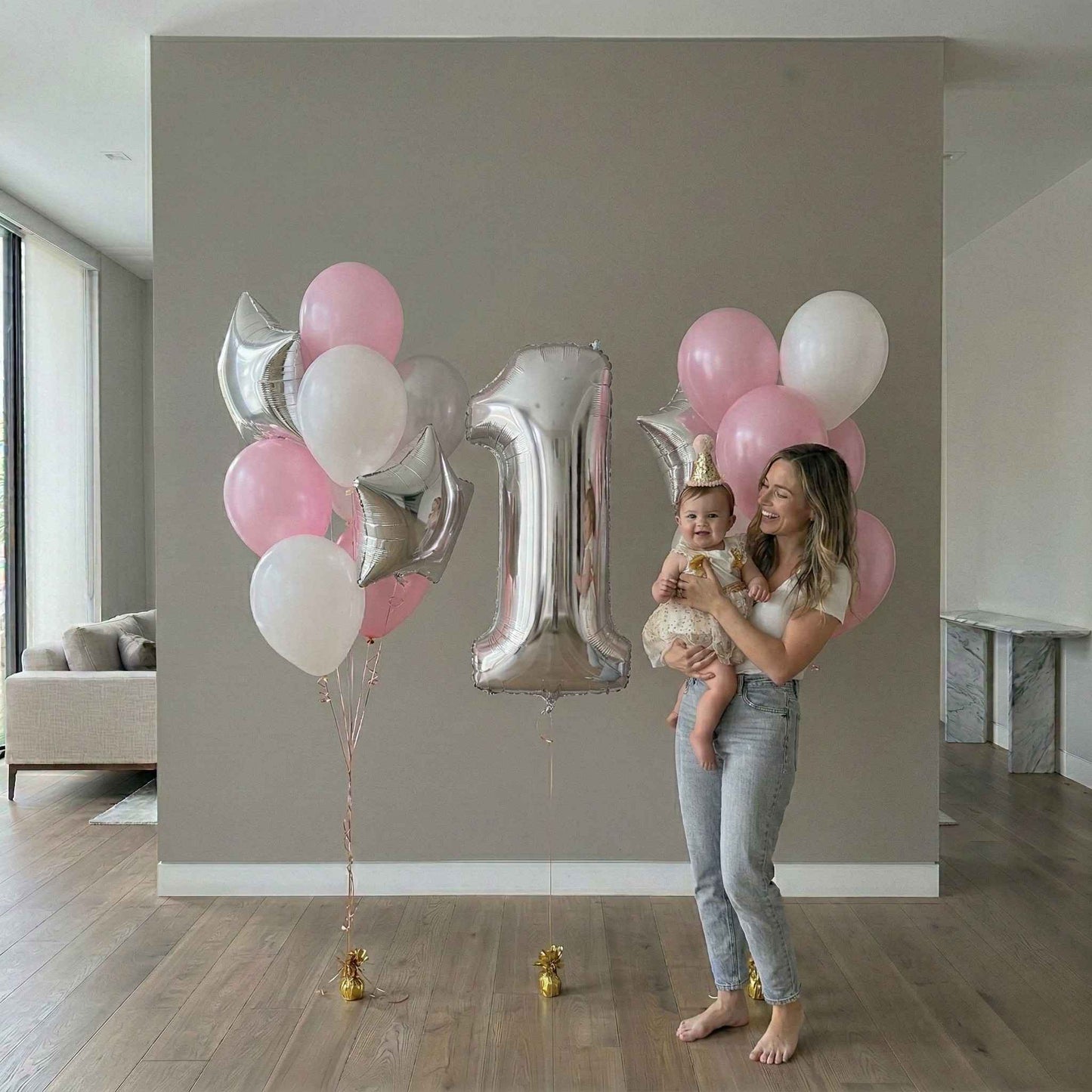 Woman holding a baby with pink, white, and silver balloons against a gray wall.