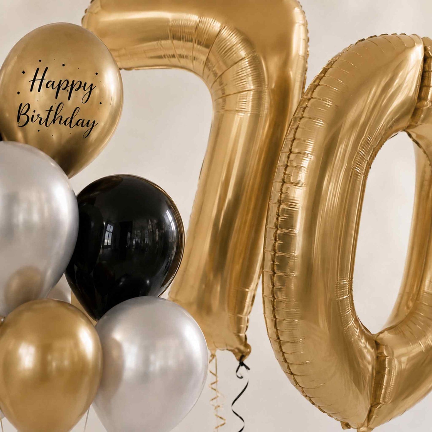Gold foil balloons spelling out '70' with a round gold balloon printed 'Happy Birthday' in black script, alongside black, silver, and gold latex balloons on a light neutral background.