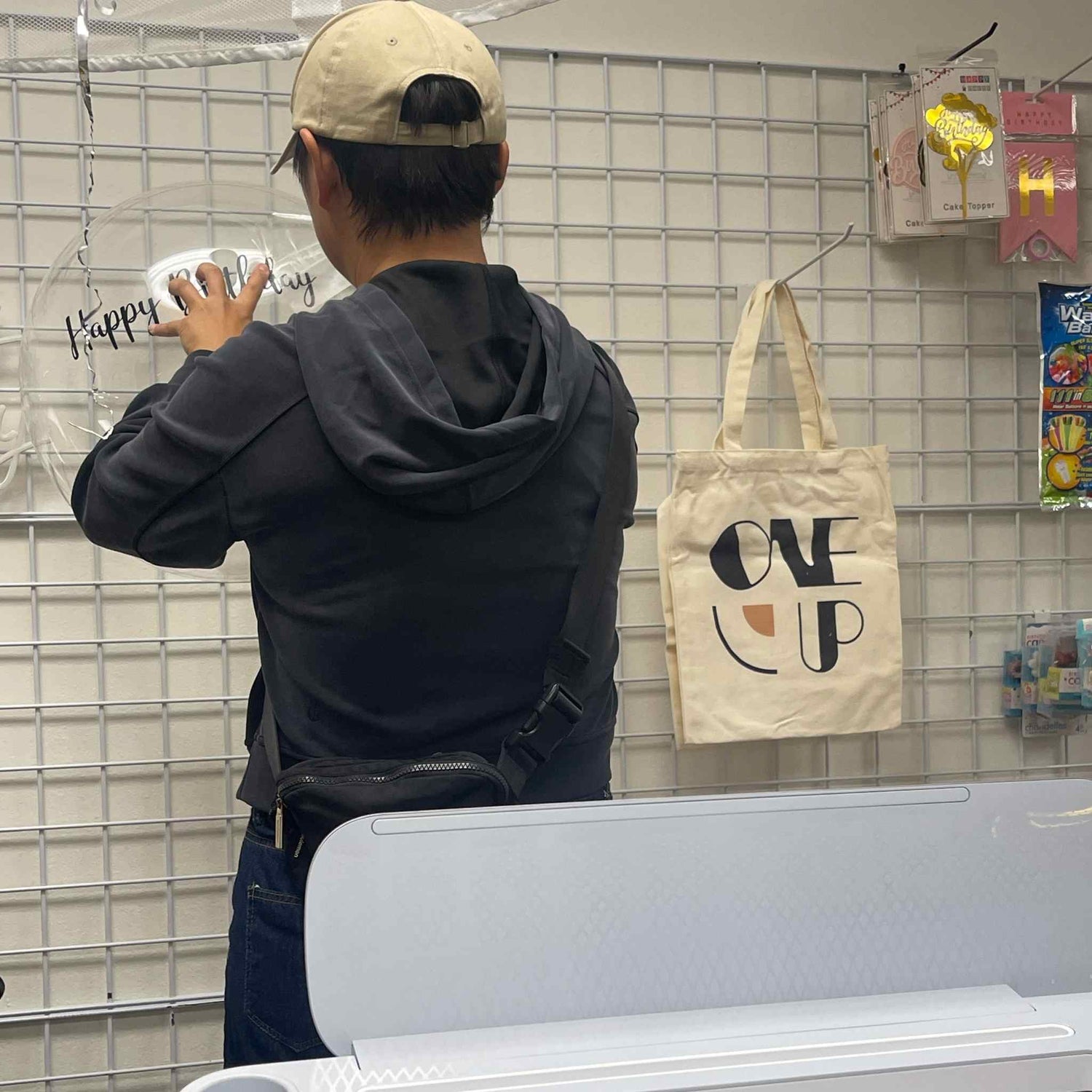 Person holding a cup with a 'Happy' design, standing next to a tote bag with 'ONE UP' text in a store setting.