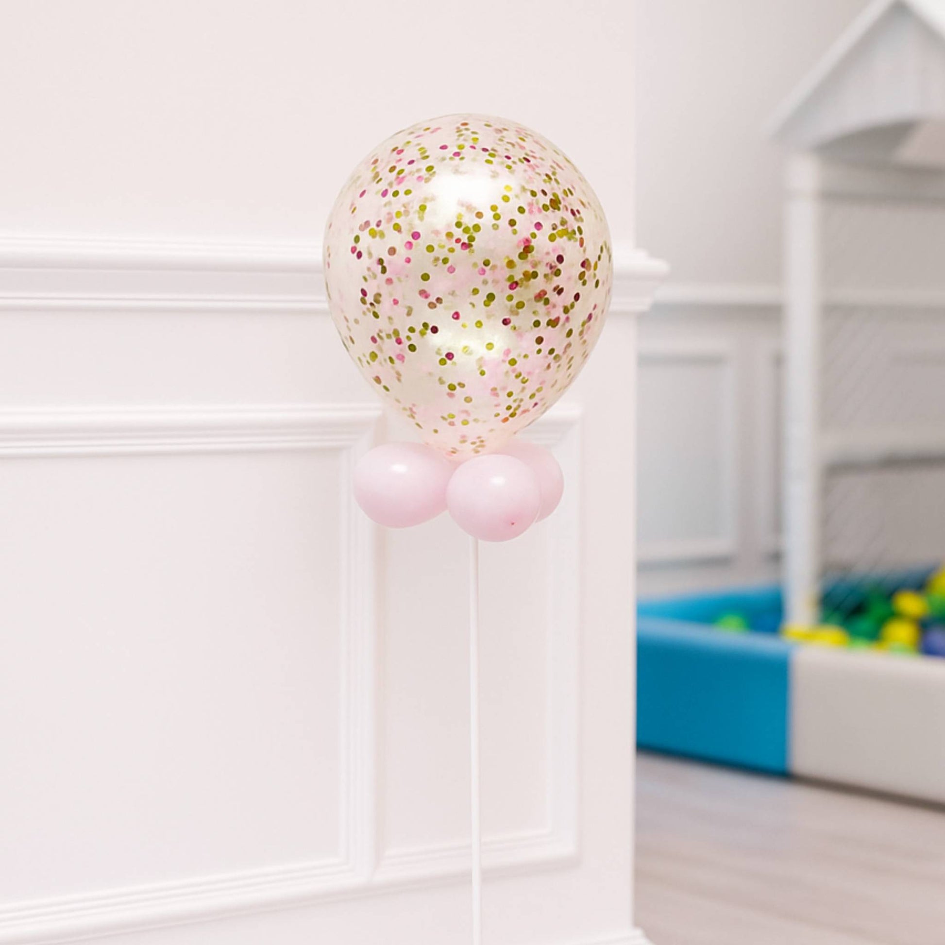 Balloon with confetti and pink base in a room with a white wall and colorful toy box.