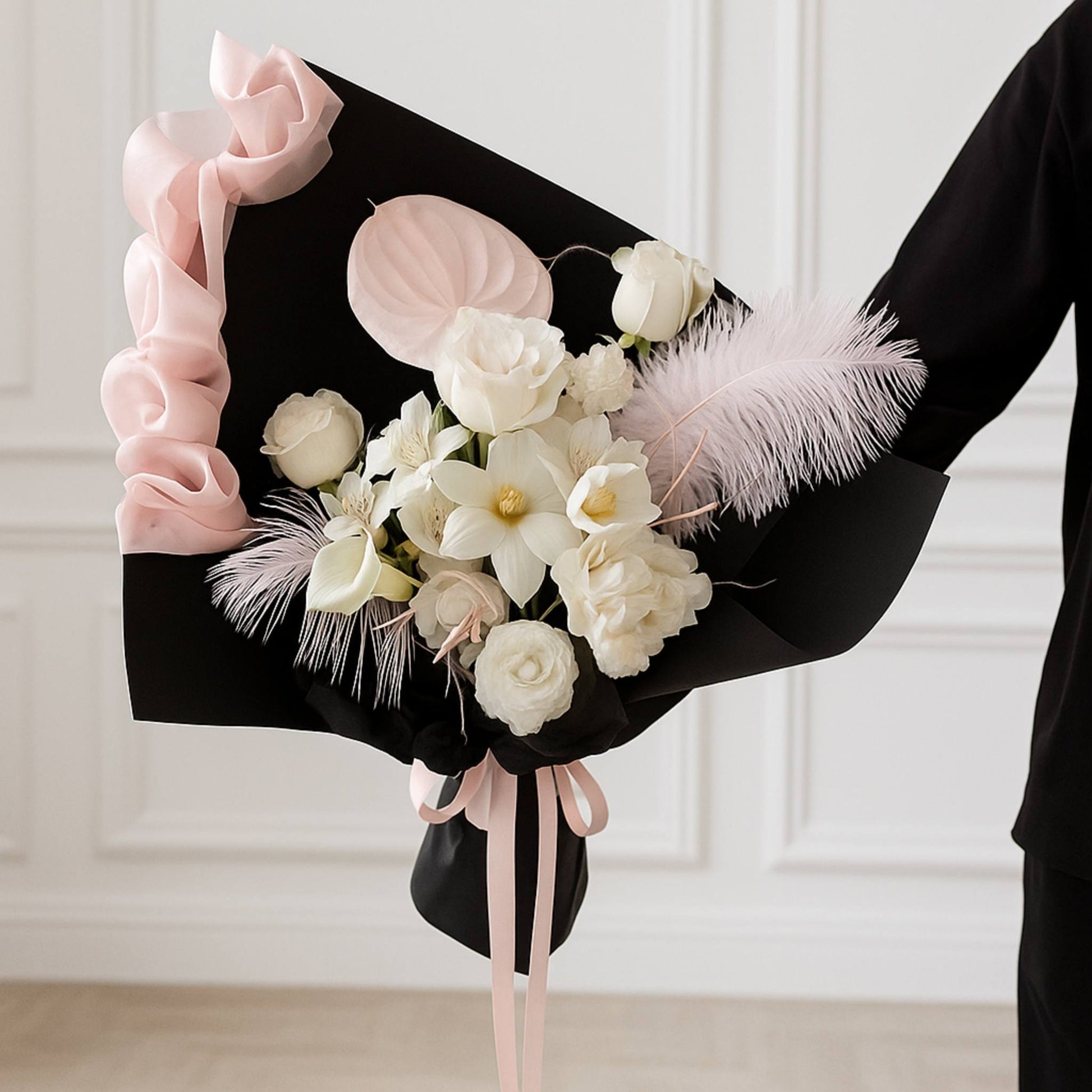 Bouquet of white flowers with pink and black accents held by a person against a white wall.