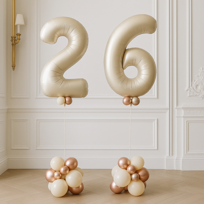 Jumbo gold helium balloons shaped as the number 26 with clusters of smaller gold and white balloons at the base, displayed in a room with white panel walls.