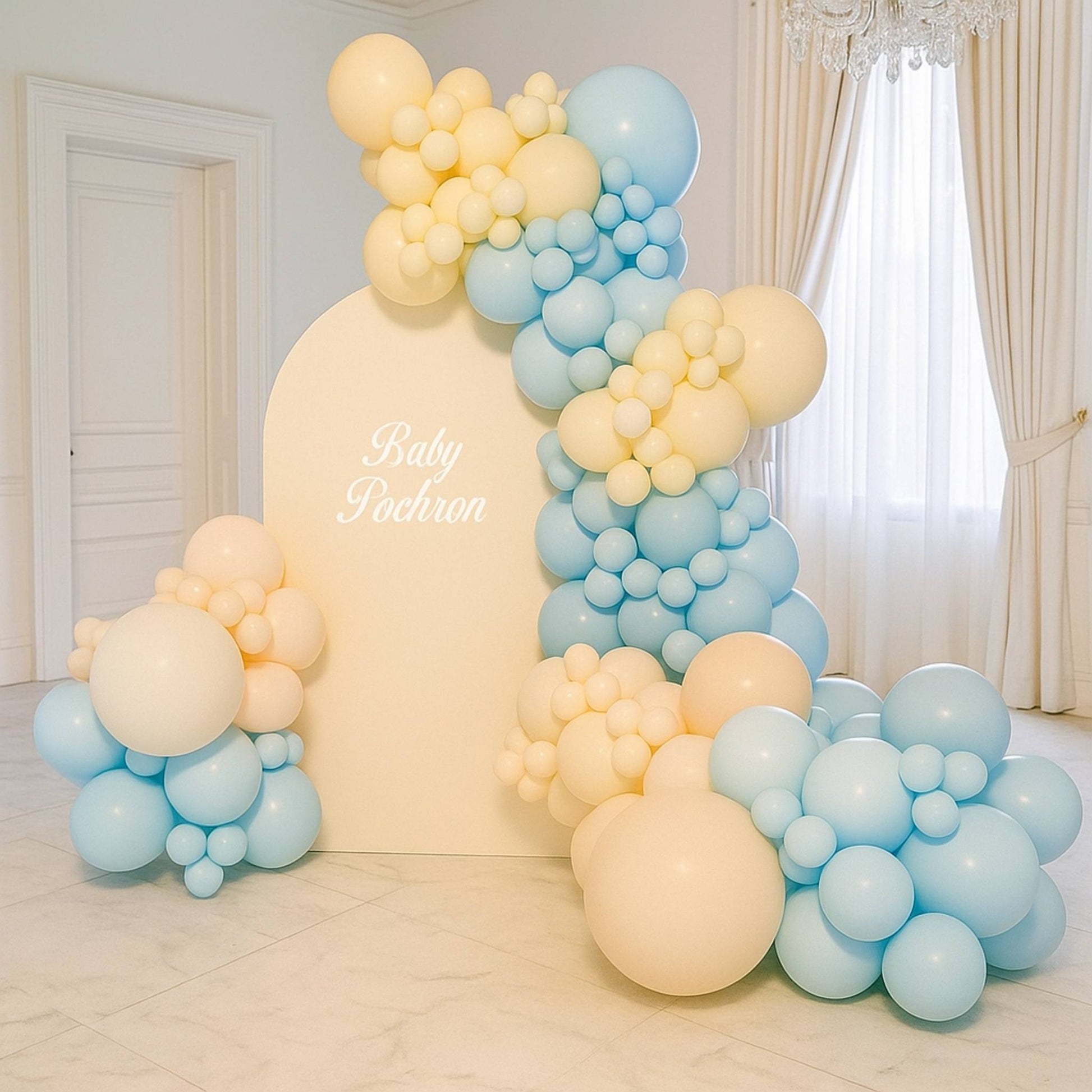 Balloon arch with blue and yellow balloons in a room with white walls and curtains.