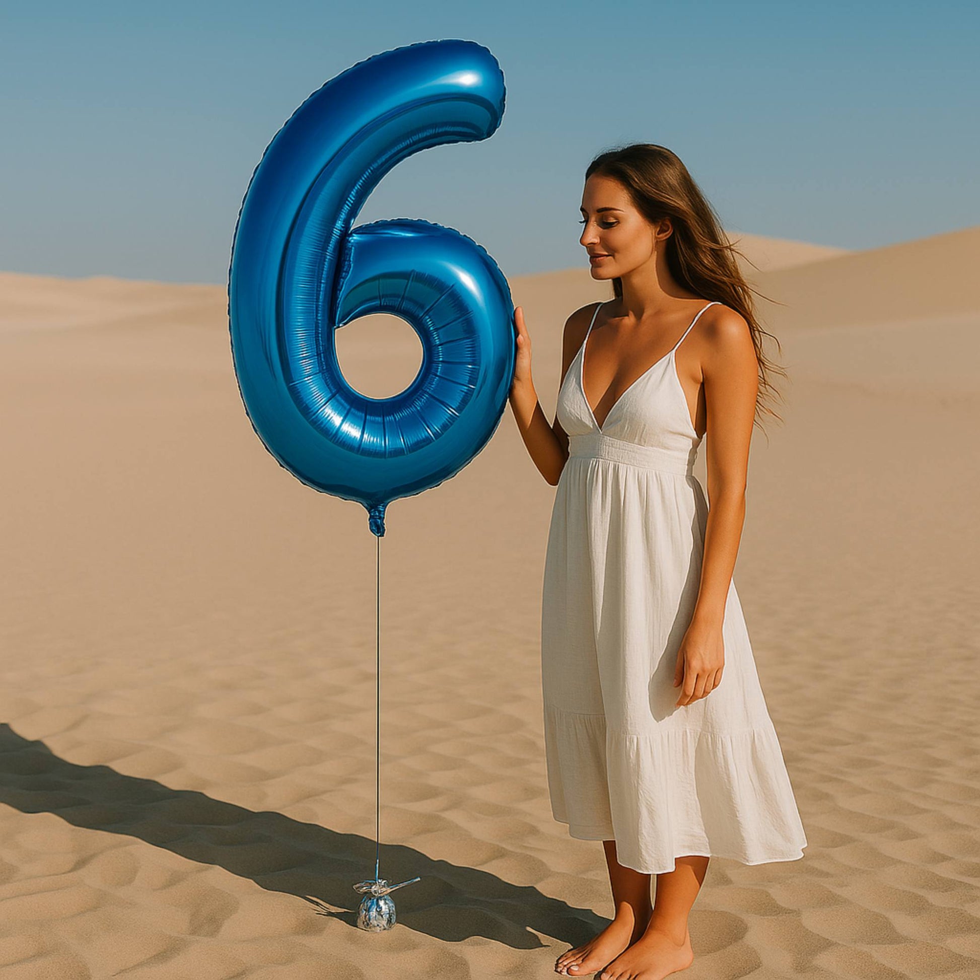 Woman in a white dress holding a large blue number 6 balloon in the desert.