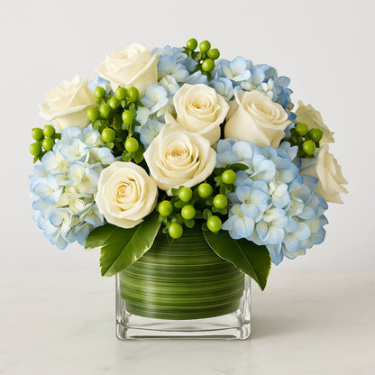 Floral arrangement with white roses, blue hydrangeas, and green berries in a clear vase on a light background