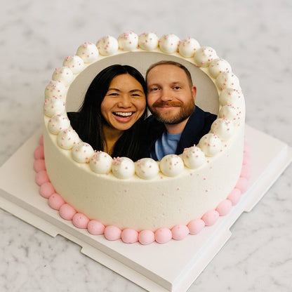 Heart-shaped cake with a photo of two people in the center on a marble surface.