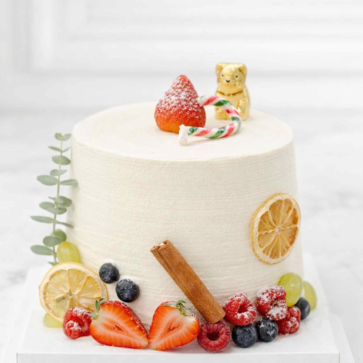 Decorative cake with fruits, a cinnamon stick, and a teddy bear on top against a white background